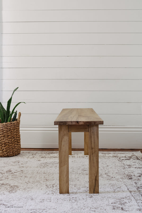Side image of the wooden bench in front of the white wall, with some greenery on the side.