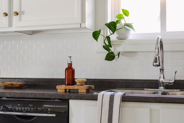 Wooden pedestal stand with a bottle brush and soap dispenser set on it sitting beside a kitchen sink.