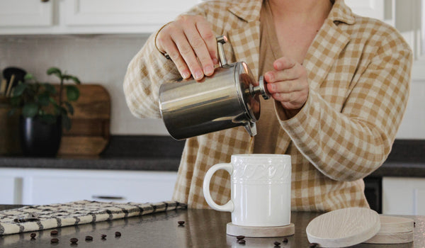 Four wooden coaster sitting on kitchen counter. One of the coasters  has a coffee mug sitting on it. There is a person pouring coffee in the cup from a French Press. 