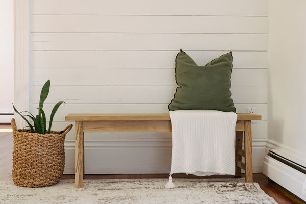 Rustic Maple bench in front of white wall. Styled with a white blanket and green throw pillow. Green plant in a woven basket sitting beside the bench.