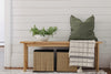 Classic bench in front of a white wall. Styled with a tan throw blanket and green pillow on one side of the bench with some greenery in a white bowl on the other side.