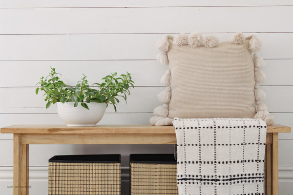 Classic maple bench styled against a white wall. Styled with a tan throw blanket and a tan pillow on one end and some greenery on the other end of the bench.